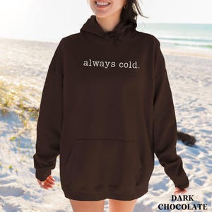 May include: Dark chocolate hoodie with the text "always cold" in white. The hoodie has a front pocket and a hood. The image is taken on a beach with the ocean in the background.