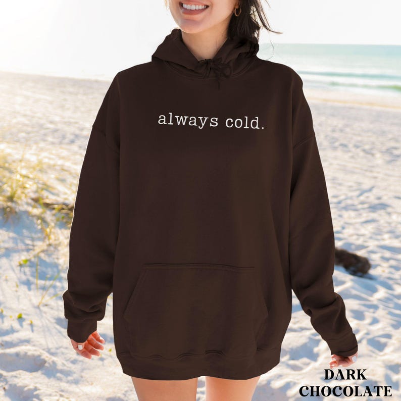 May include: Dark chocolate hoodie with the text "always cold" in white. The hoodie has a front pocket and a hood. The image is taken on a beach with the ocean in the background.