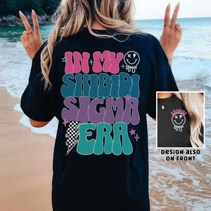 Puede incluir: Camiseta negra con un gr&aacute;fico colorido que dice "In My Shibidi Sigma Era" con una cara sonriente que gotea. El dise&ntilde;o tambi&eacute;n est&aacute; en la parte delantera de la camiseta.