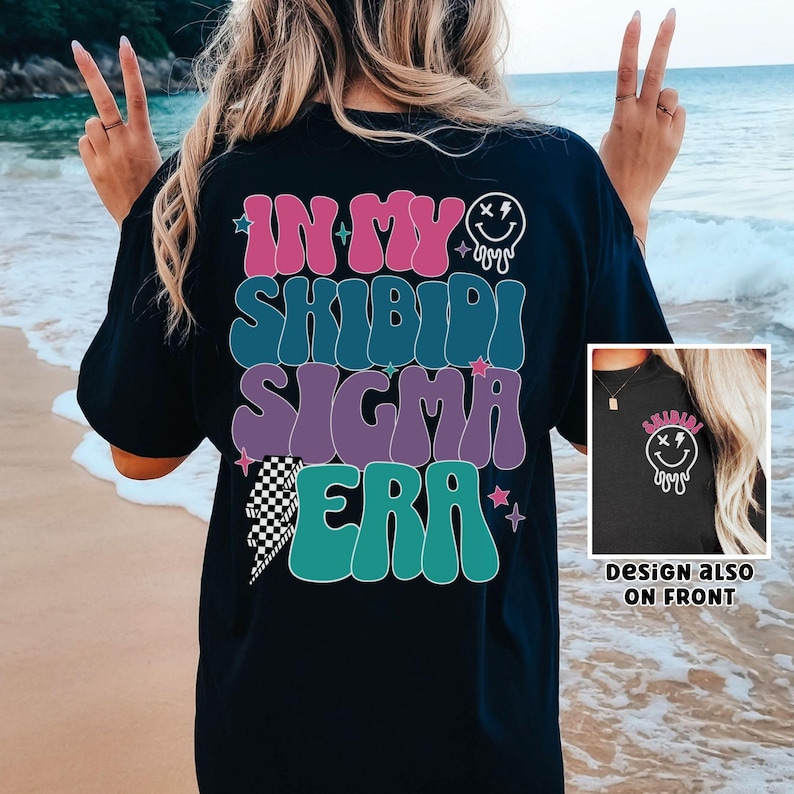 Puede incluir: Camiseta negra con un gr&aacute;fico colorido que dice "In My Shibidi Sigma Era" con una cara sonriente que gotea. El dise&ntilde;o tambi&eacute;n est&aacute; en la parte delantera de la camiseta.