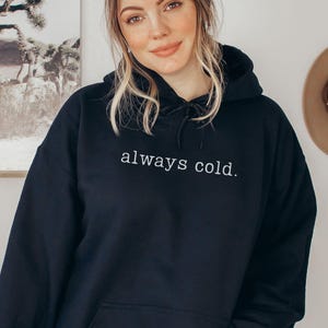 May include: Black hooded sweatshirt with the text "always cold." The sweatshirt has a drawstring hood and a front pocket. The text is in a simple, white sans-serif font.