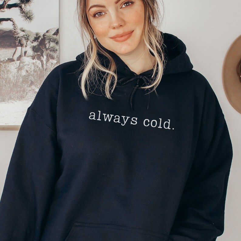 May include: Black hooded sweatshirt with the text "always cold." The sweatshirt has a drawstring hood and a front pocket. The text is in a simple, white sans-serif font.