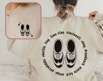 May include: A sand-colored crewneck sweatshirt featuring a black graphic of tap shoes on the back, surrounded by the text: flap, time step, cincinnati, chug, drawback, maxie ford, wings, paradiddle, shuffle. The front chest has a smaller tap shoe graphic.