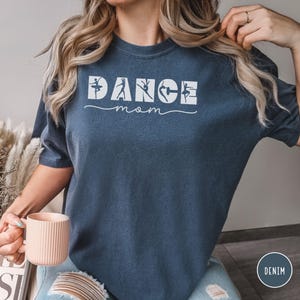 May include: A denim blue short-sleeved t-shirt featuring the white text DANCE with silhouettes of dancers integrated into the letters, and the word mom written in a cursive script underneath. The shirt has a crew neckline and a relaxed fit.