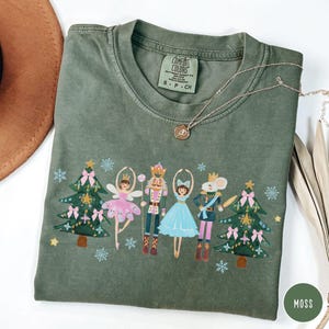 May include: A moss green crew neck t-shirt featuring a graphic print of two Christmas trees, a fairy, a nutcracker, a dancer, and a mouse soldier. The shirt has a Comfort Colors label at the neck and is paired with a gold-toned pendant necklace.