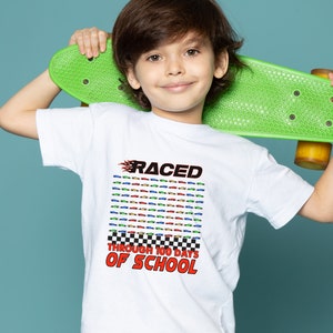 K&ouml;nnte beinhalten: Wei&szlig;es T-Shirt mit dem Text "RACED THROUGH 100 DAYS OF SCHOOL" in Rot und Schwarz. Das Shirt zeigt ein Raster aus bunten Rennwagen. Das Kind h&auml;lt ein gr&uuml;nes Skateboard.