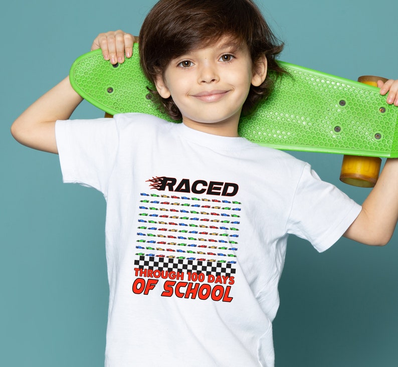 K&ouml;nnte beinhalten: Wei&szlig;es T-Shirt mit dem Text "RACED THROUGH 100 DAYS OF SCHOOL" in Rot und Schwarz. Das Shirt zeigt ein Raster aus bunten Rennwagen. Das Kind h&auml;lt ein gr&uuml;nes Skateboard.