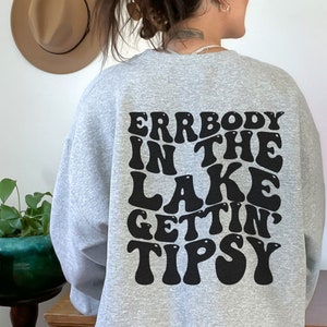 May include: A light gray sweatshirt with the text "ERRBODY IN THE LAKE GETTIN TIPSY" in a black, retro font. The sweatshirt is a casual style, perfect for a relaxed day.
