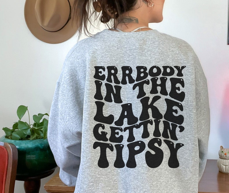 May include: A light gray sweatshirt with the text "ERRBODY IN THE LAKE GETTIN TIPSY" in a black, retro font. The sweatshirt is a casual style, perfect for a relaxed day.