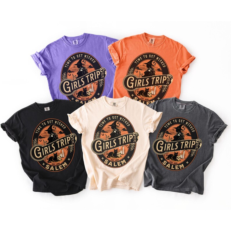 May include: Five short-sleeve t-shirts in purple, orange, black, cream, and dark gray. Each shirt features a circular graphic with a witch, a black cat, and the text: TIME TO GET WICKED, GIRLS TRIP, SALEM, EST 1692, 2026.