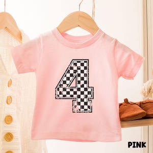 Pu&ograve; includere: Una maglietta rosa chiaro per bambini con il numero "4" in un motivo a scacchi bianco e nero. La parola "PINK" &egrave; stampata in nero in basso a destra. La maglietta &egrave; appesa a un appendiabiti di legno.