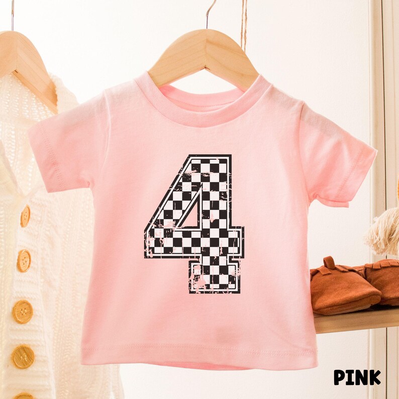 Pu&ograve; includere: Una maglietta rosa chiaro per bambini con il numero "4" in un motivo a scacchi bianco e nero. La parola "PINK" &egrave; stampata in nero in basso a destra. La maglietta &egrave; appesa a un appendiabiti di legno.