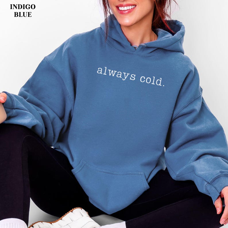 May include: Indigo blue hoodie with the text "always cold" in white. The hoodie has a front pocket and a drawstring hood. The model is wearing black leggings and white sneakers.