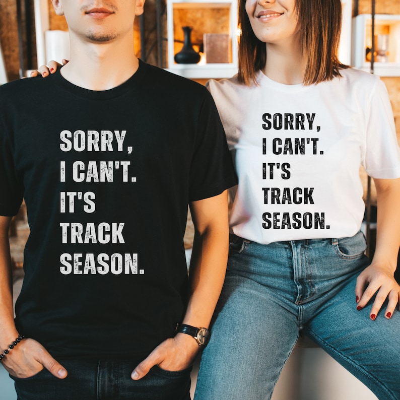 May include: Two black and white t-shirts with the text "Sorry, I can't. It's track season." on them. The shirts are worn by two people standing next to each other.
