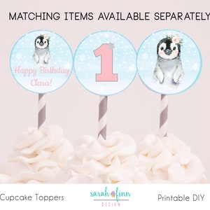 May include: Three cupcake toppers featuring a penguin, a number one, and a penguin with the text "Happy Birthday Clara!" on a blue background with white snowflakes. The toppers are on white frosting.