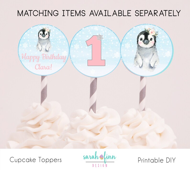 May include: Three cupcake toppers featuring a penguin, a number one, and a penguin with the text "Happy Birthday Clara!" on a blue background with white snowflakes. The toppers are on white frosting.