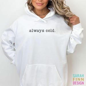 May include: White hoodie with a front pocket and drawstring hood. The text "always cold." is printed in black across the chest. The Sarah Finn Design logo is visible in the bottom right corner.
