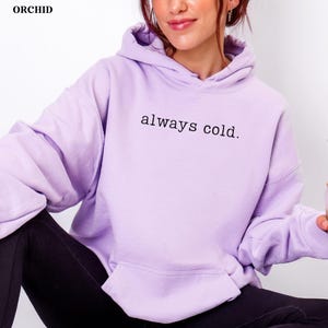 May include: A light purple hoodie with the text "always cold." printed in black. The hoodie has a hood and a front pocket. The brand name "ORCHID" is in the top left corner.