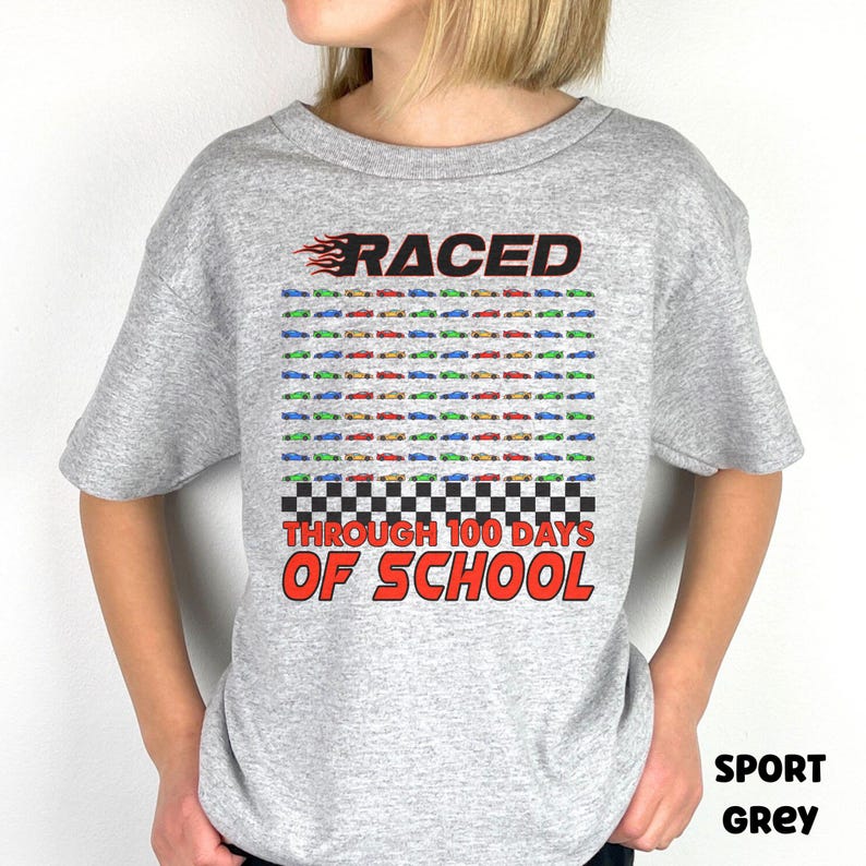 K&ouml;nnte beinhalten: Hellgraues T-Shirt mit dem Text "RACED THROUGH 100 DAYS OF SCHOOL" in Rot und Schwarz. Das Design umfasst Reihen farbiger Rennwagen und ein Schachbrettmuster.
