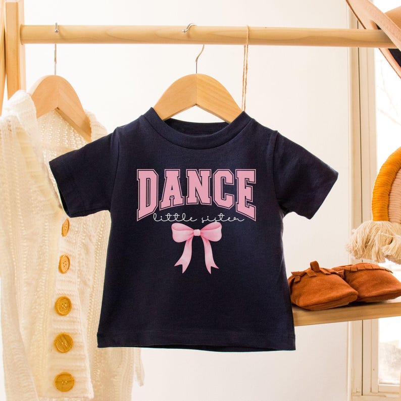 K&ouml;nnte beinhalten: Ein marineblaues T-Shirt mit dem Text "DANCE little sister" in rosa Schrift und einer rosa Schleife.