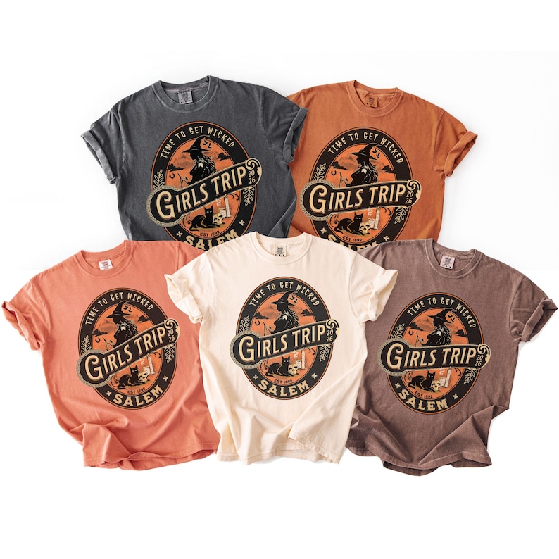 May include: Five short-sleeved crew neck t-shirts in charcoal, orange, peach, cream, and brown. Each shirt features a circular graphic with a witch, a black cat, and the text "TIME TO GET WICKED", "GIRLS TRIP", "SALEM", and "2026" in a vintage style.