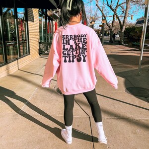 May include: A light pink sweatshirt with the text "ERRBODY IN THE LAKE GETTIN' TIPSY" in black. The sweatshirt is paired with black leggings and white sneakers. The person is standing on a sidewalk in front of a building.