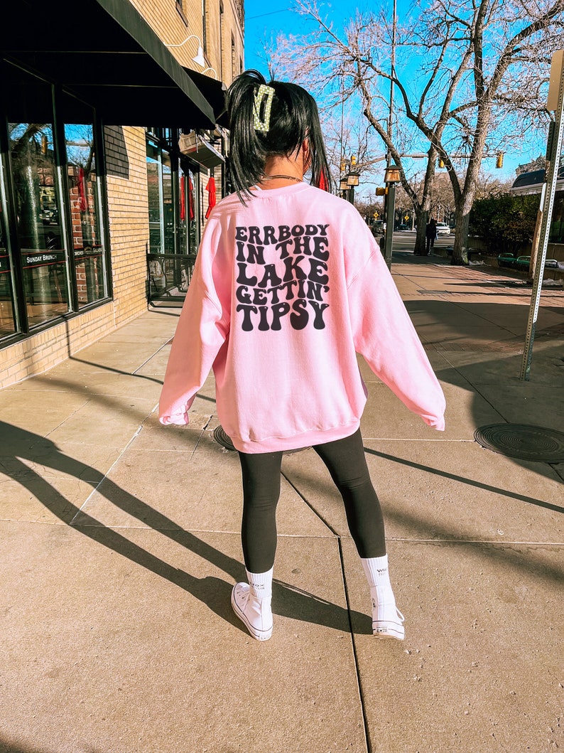 May include: A light pink sweatshirt with the text "ERRBODY IN THE LAKE GETTIN' TIPSY" in black. The sweatshirt is paired with black leggings and white sneakers. The person is standing on a sidewalk in front of a building.