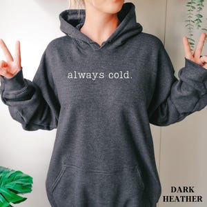 May include: Dark heather gray hoodie with the text "always cold." printed in white. The hoodie has a drawstring hood and a front pocket. The words "Dark Heather" are printed in the bottom right corner.