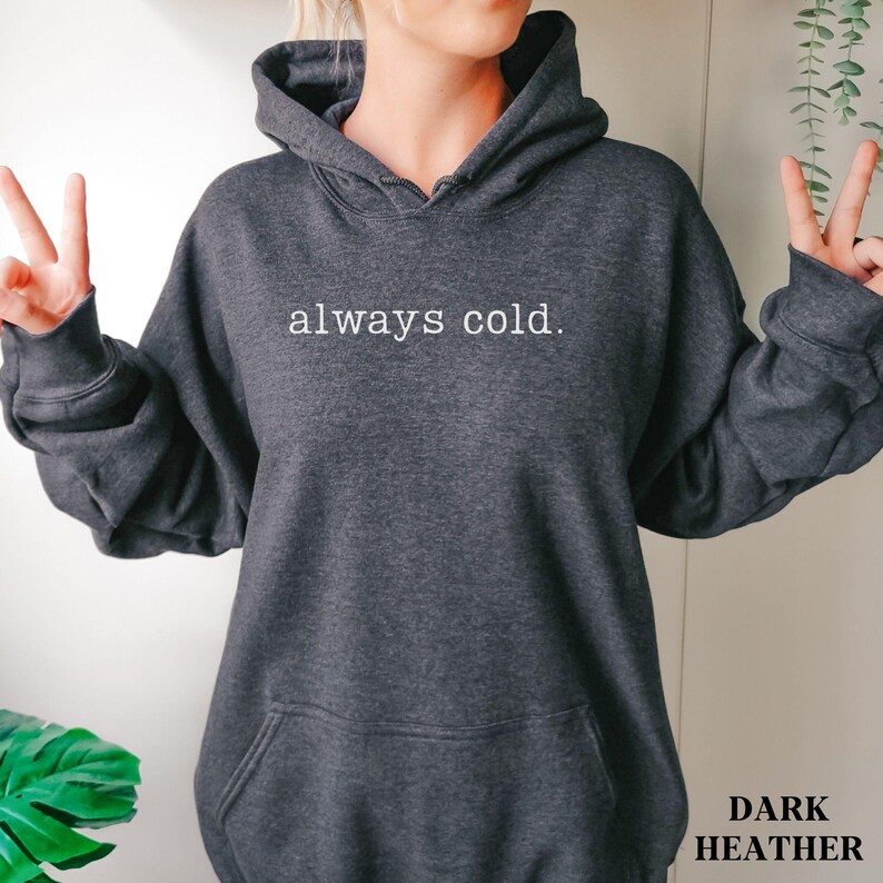 May include: Dark heather gray hoodie with the text "always cold." printed in white. The hoodie has a drawstring hood and a front pocket. The words "Dark Heather" are printed in the bottom right corner.