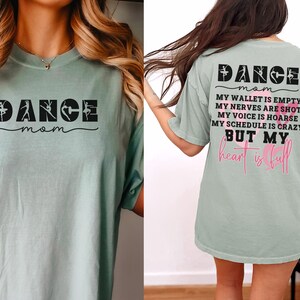 May include: A sage green t-shirt featuring black text on the front that reads DANCE mom with dancer silhouettes. The back displays the text DANCE mom, MY WALLET IS EMPTY, MY NERVES ARE SHOT, MY VOICE IS HOARSE, MY SCHEDULE IS CRAZY BUT MY heart is full.