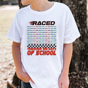 K&ouml;nnte beinhalten: Wei&szlig;es T-Shirt mit farbenfroher Grafik. Das Design zeigt das Wort "RACED" in Rot mit Flammen, ein Raster von Spielzeugautos und den Text "THROUGH 100 DAYS OF SCHOOL" in Rot und Schwarz.