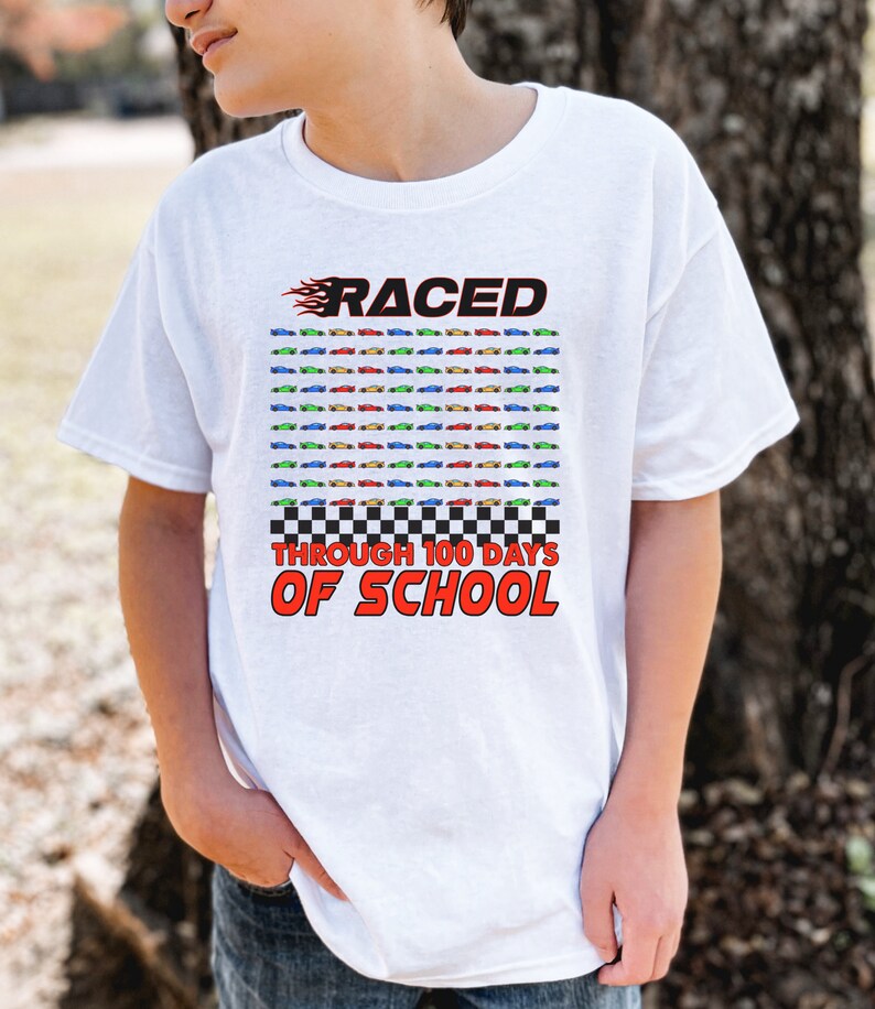K&ouml;nnte beinhalten: Wei&szlig;es T-Shirt mit farbenfroher Grafik. Das Design zeigt das Wort "RACED" in Rot mit Flammen, ein Raster von Spielzeugautos und den Text "THROUGH 100 DAYS OF SCHOOL" in Rot und Schwarz.