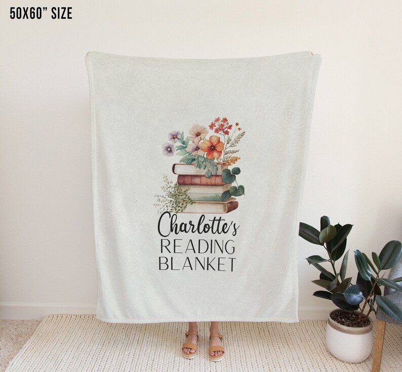 May include: A white fleece blanket with a floral design and a stack of books. The text "Charlotte's Reading Blanket" is printed on the blanket.