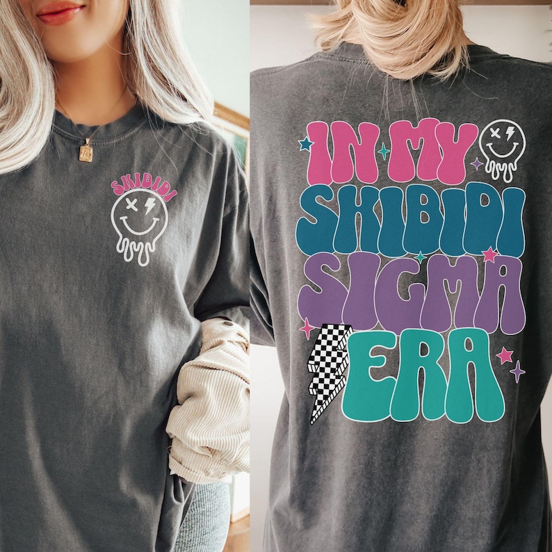 Puede incluir: Una camiseta de manga larga gris oscuro con un estampado gr&aacute;fico en la parte delantera y trasera. El gr&aacute;fico delantero es una cara sonriente blanca con un rayo y l&iacute;neas que gotean. El gr&aacute;fico trasero es un dise&ntilde;o de texto multicolor que dice "In My Shibidi Sigma Era" con un rayo y estrellas.
