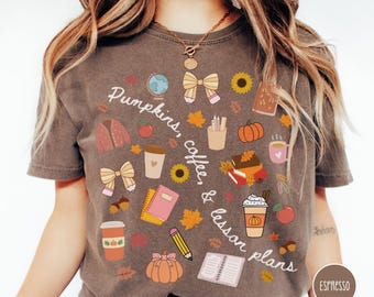 May include: A dark brown t-shirt featuring a graphic print with the text "Pumpkins, coffee, & lesson plans" surrounded by illustrations of a globe, bows, sunflowers, books, notebooks, pencils, coffee cups, apples, acorns, leaves, and pumpkins.