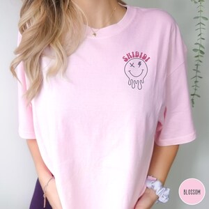 Puede incluir: Camiseta rosa con un gr&aacute;fico de una cara sonriente que gotea con una X para un ojo y un rayo para el otro. El texto "SKIBIDI" est&aacute; por encima de la cara sonriente.