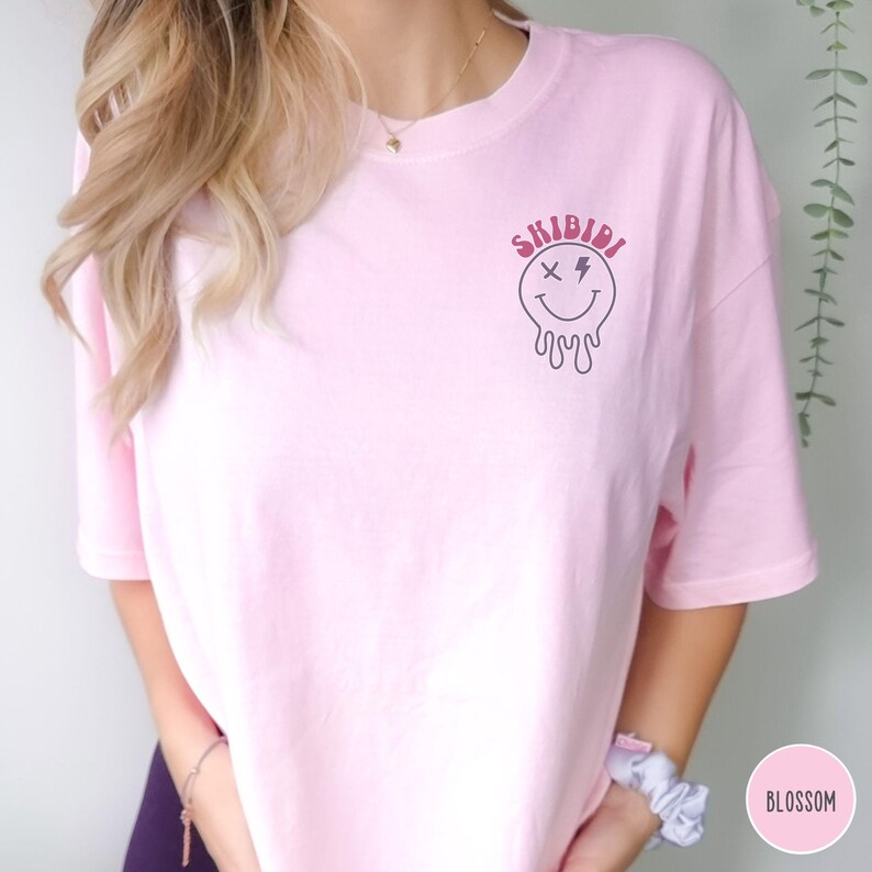 Puede incluir: Camiseta rosa con un gr&aacute;fico de una cara sonriente que gotea con una X para un ojo y un rayo para el otro. El texto "SKIBIDI" est&aacute; por encima de la cara sonriente.