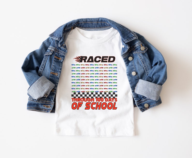 K&ouml;nnte beinhalten: Wei&szlig;es T-Shirt mit dem Text "RACED THROUGH 100 DAYS OF SCHOOL" in Rot und Schwarz. Das Shirt zeigt ein Raster aus bunten Rennwagen und wird auf einer Jeansjacke pr&auml;sentiert.