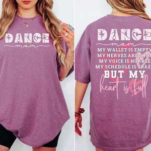 May include: A berry-colored short-sleeve t-shirt featuring the white text DANCE MOM on the front. The back displays the text DANCE MOM, MY WALLET IS EMPTY, MY NERVES ARE SHOT, MY VOICE IS HOARSE, MY SCHEDULE IS CRAZY BUT MY heart is full with hearts.
