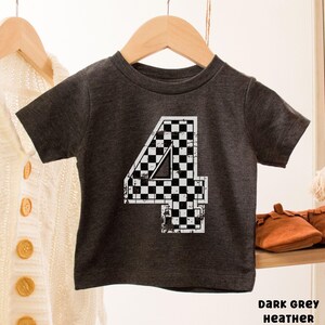 Pu&ograve; includere: T-shirt da bambino in grigio scuro m&eacute;lange con un grande numero 4 a scacchi bianchi e neri. Il numero ha un aspetto invecchiato e vintage. La maglietta &egrave; appesa a un appendiabiti in legno.