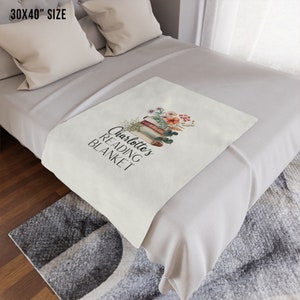 May include: A white throw blanket with a floral design and the text "Charlotte's Reading Blanket" printed on it. The blanket is folded and placed on a bed with white pillows.