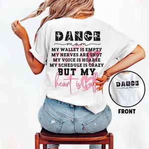 May include: A white t-shirt featuring black text that reads DANCE MOM, MY WALLET IS EMPTY, MY NERVES ARE SHOT, MY VOICE IS HOARSE, MY SCHEDULE IS CRAZY, BUT MY heart is full, with pink heart outlines and a small circular front view inset.