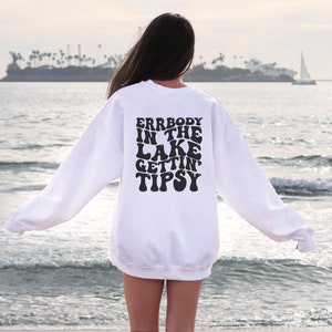 May include: A white sweatshirt with the text "ERRBODY IN THE LAKE GETTIN' TIPSY" in black, retro-style lettering. The sweatshirt is seen from the back, with a beach and ocean in the background.