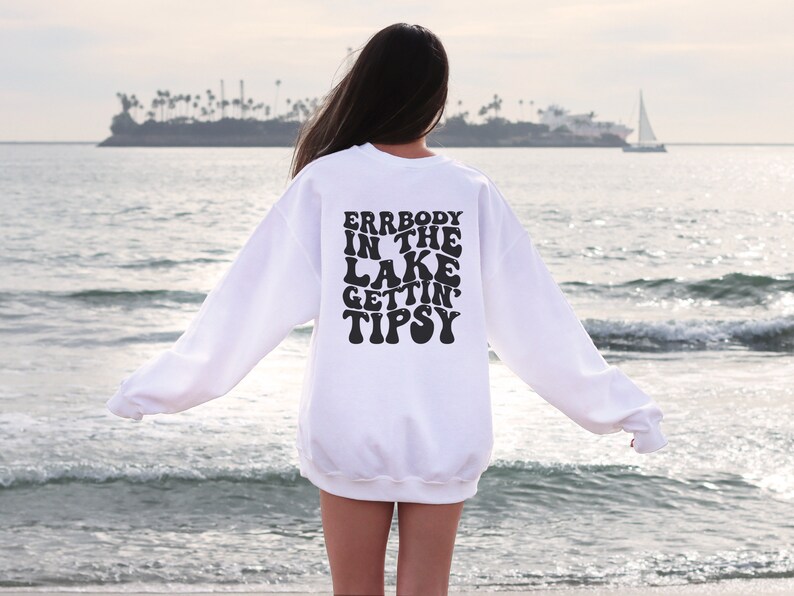 May include: A white sweatshirt with the text "ERRBODY IN THE LAKE GETTIN' TIPSY" in black, retro-style lettering. The sweatshirt is seen from the back, with a beach and ocean in the background.