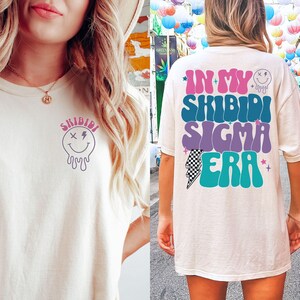 Puede incluir: Dos camisetas blancas con un dise&ntilde;o gr&aacute;fico. La parte delantera de la camiseta tiene una cara sonriente que gotea con el texto "Shibidi". La parte trasera de la camiseta tiene el texto "In My Shibidi Sigma Era" en una fuente retro con un gr&aacute;fico de rayo.