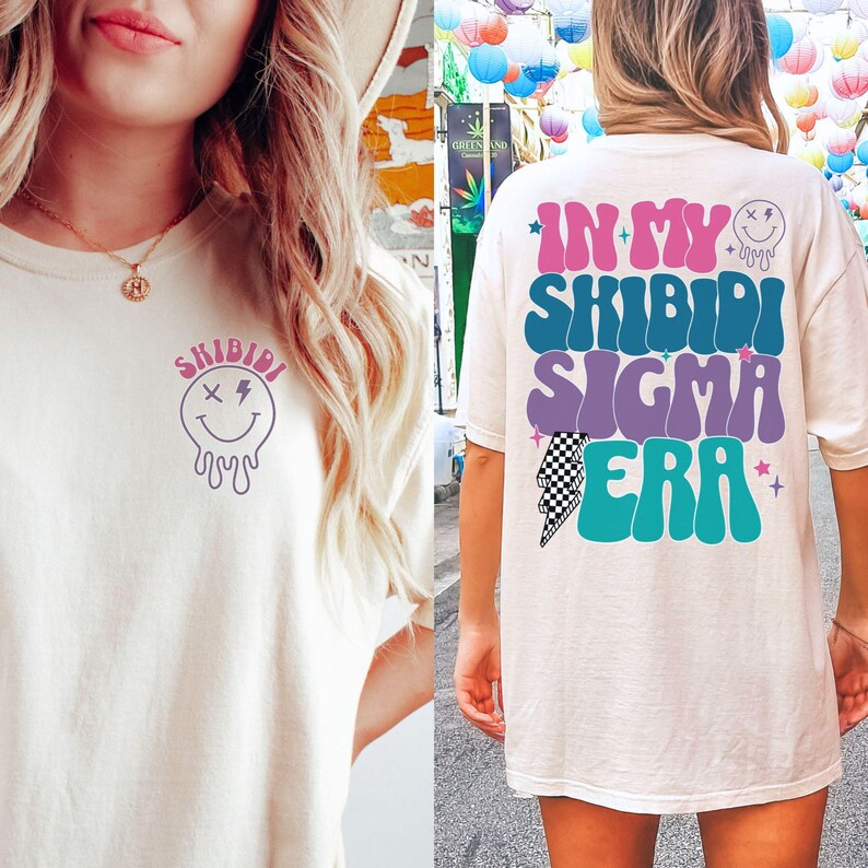 Puede incluir: Dos camisetas blancas con un dise&ntilde;o gr&aacute;fico. La parte delantera de la camiseta tiene una cara sonriente que gotea con el texto "Shibidi". La parte trasera de la camiseta tiene el texto "In My Shibidi Sigma Era" en una fuente retro con un gr&aacute;fico de rayo.