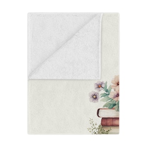 May include: A white throw blanket with a floral design and a stack of books. The flowers are pink, purple, and green. The books are brown and have a vintage look.