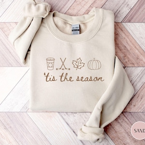 May include: A sand-colored crewneck sweatshirt featuring a brown graphic print. The design includes icons of a coffee cup, crossed hockey sticks, a maple leaf, and a pumpkin, followed by the brown text 'tis the season' in a cursive script font.