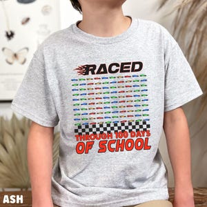 K&ouml;nnte beinhalten: Hellgraues T-Shirt mit einem bunten Rennwagen-Design. Das Shirt zeigt das Wort "RACED" &uuml;ber einem Raster von 100 kleinen Rennwagen in verschiedenen Farben, mit dem Text "THROUGH 100 DAYS OF SCHOOL" darunter.