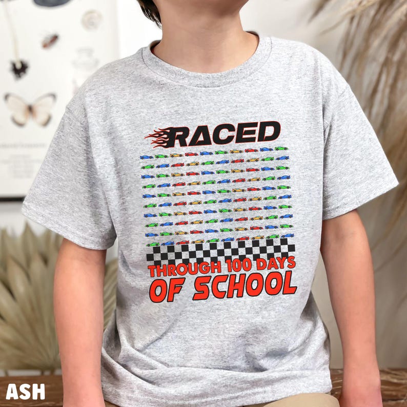 K&ouml;nnte beinhalten: Hellgraues T-Shirt mit einem bunten Rennwagen-Design. Das Shirt zeigt das Wort "RACED" &uuml;ber einem Raster von 100 kleinen Rennwagen in verschiedenen Farben, mit dem Text "THROUGH 100 DAYS OF SCHOOL" darunter.